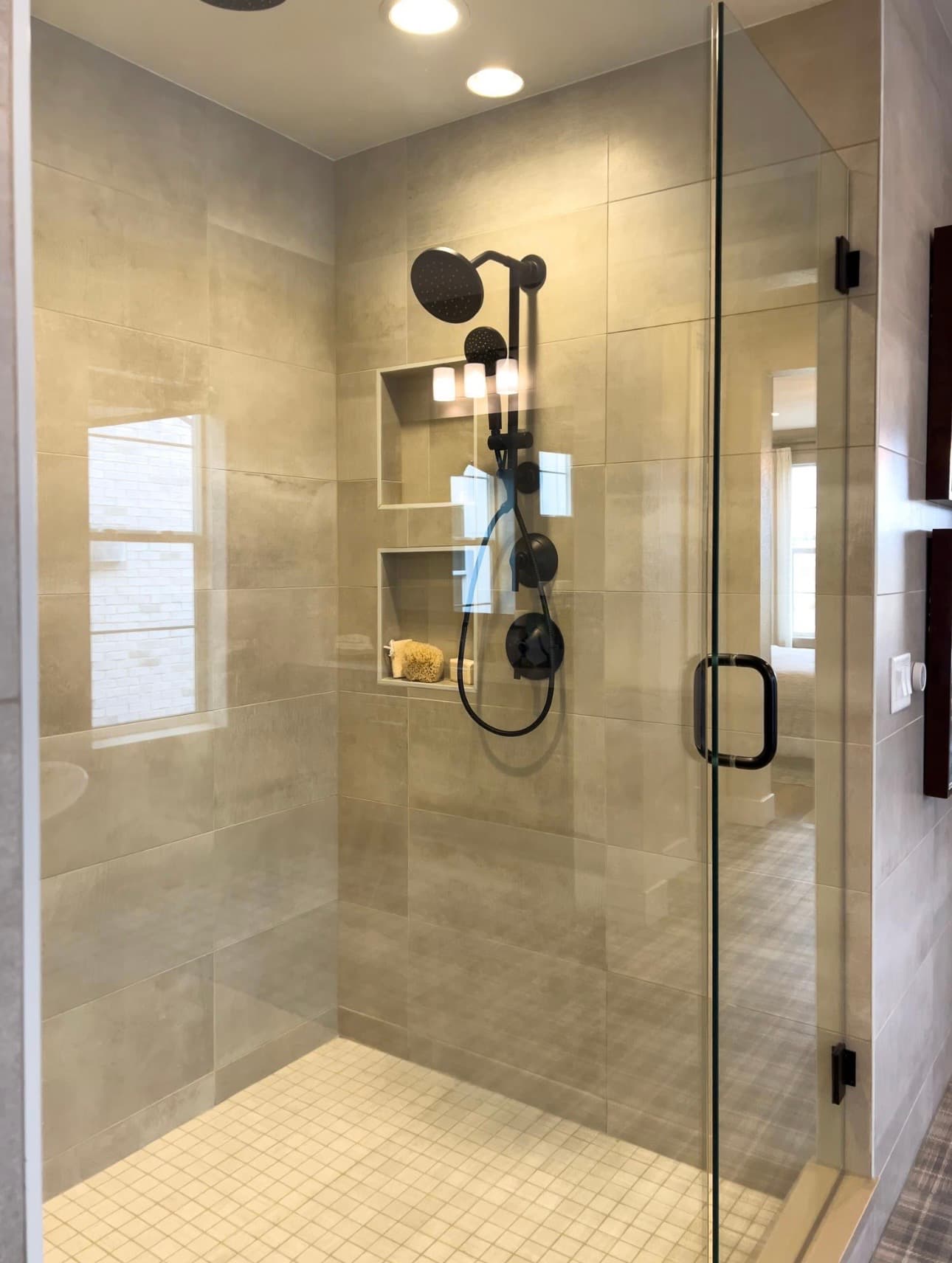Modern walk-in shower with beige tiles, black fixtures, and a glass door with black handle.