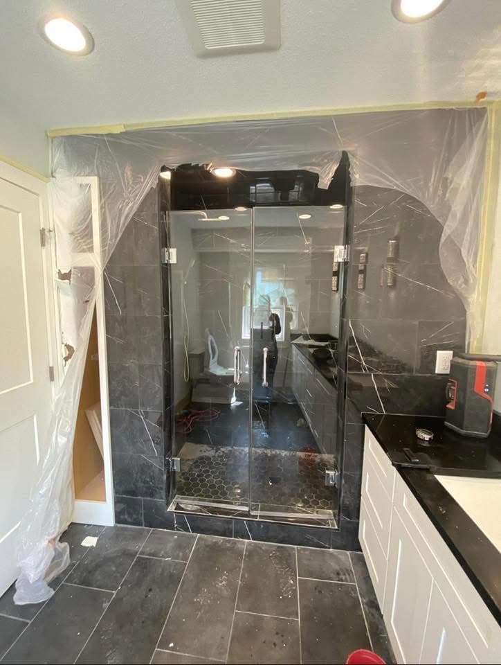 Modern bathroom renovation featuring dark marble tiles, glass shower doors, and protective plastic coverings.