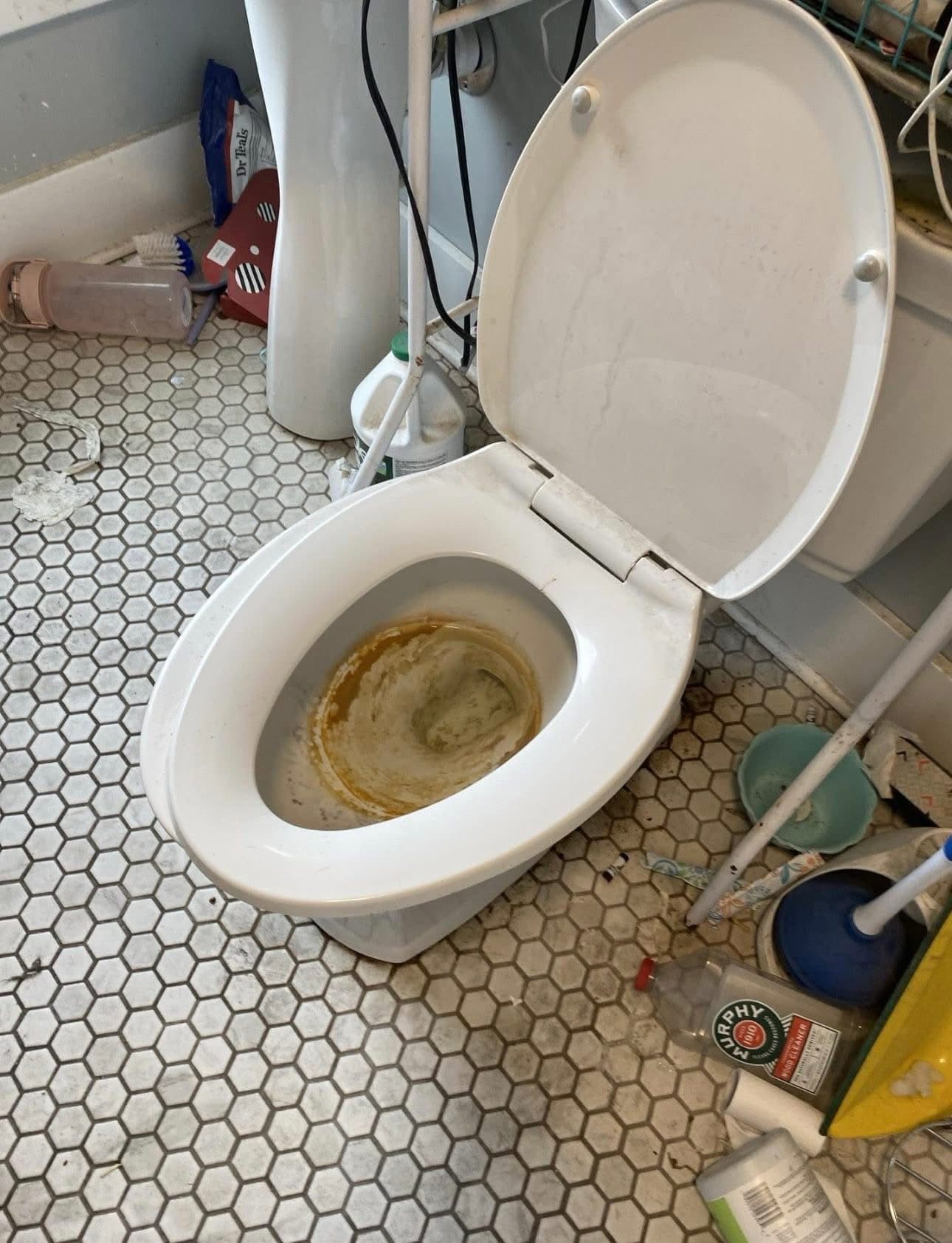 Stained white toilet with open lid on a cluttered, dirty hexagonal tile bathroom floor.