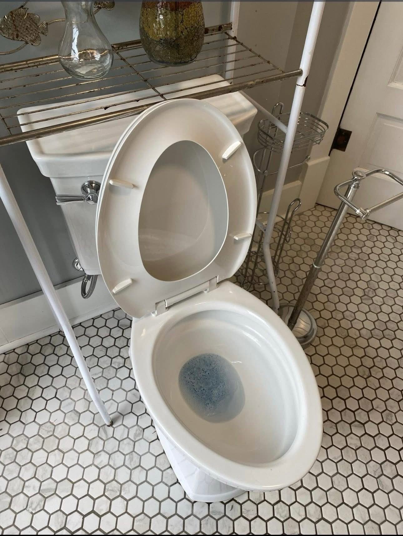 White toilet with open seat and blue water on a white hexagonal tile bathroom floor.